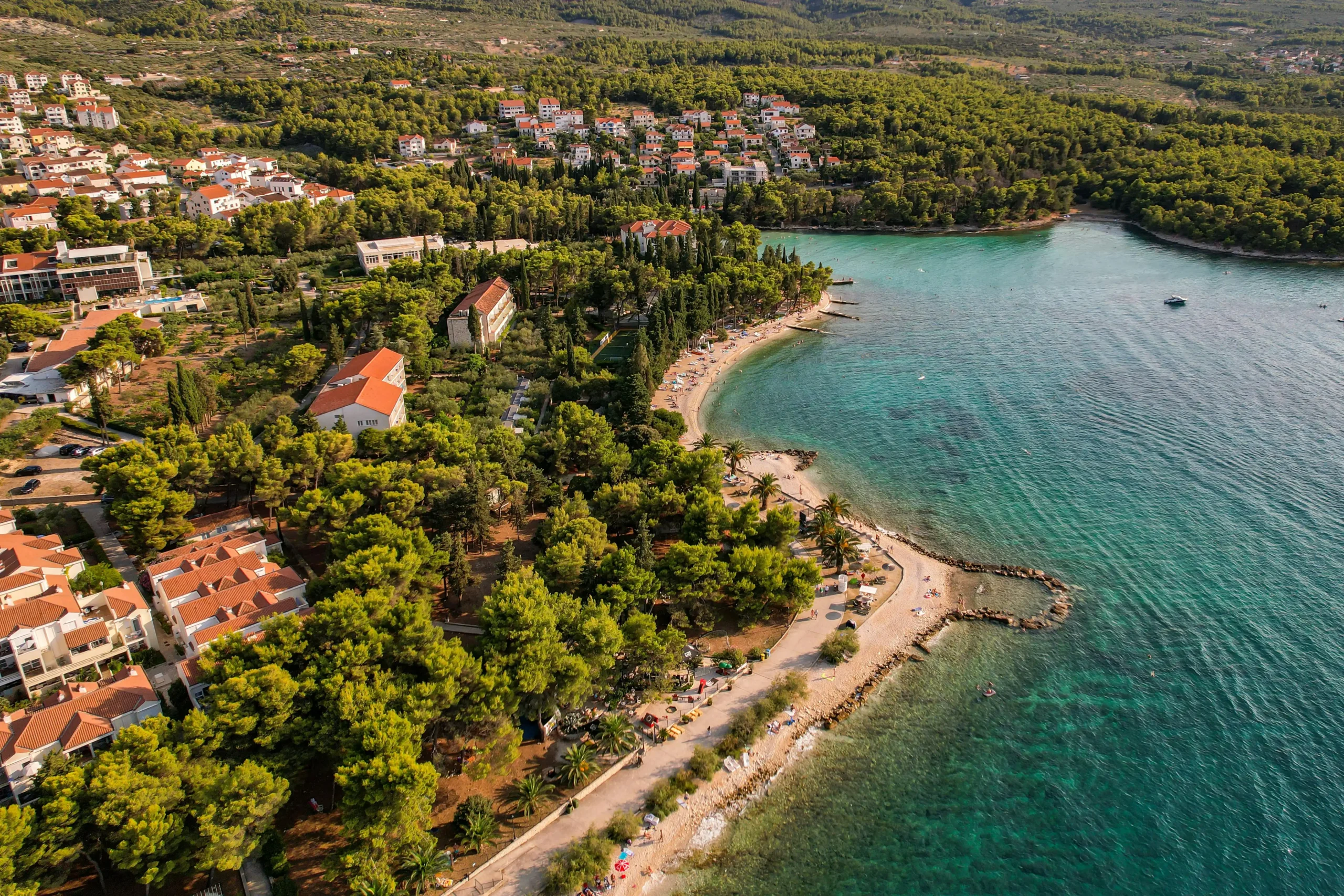 Vista aérea de un pueblo costero con playas turquesas, hoteles y pinos mediterráneos en una cala soleada.